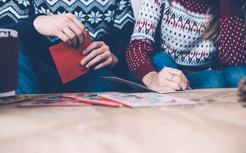 Crop shot of couple in sweaters sitting together and writing seasonal greetings.